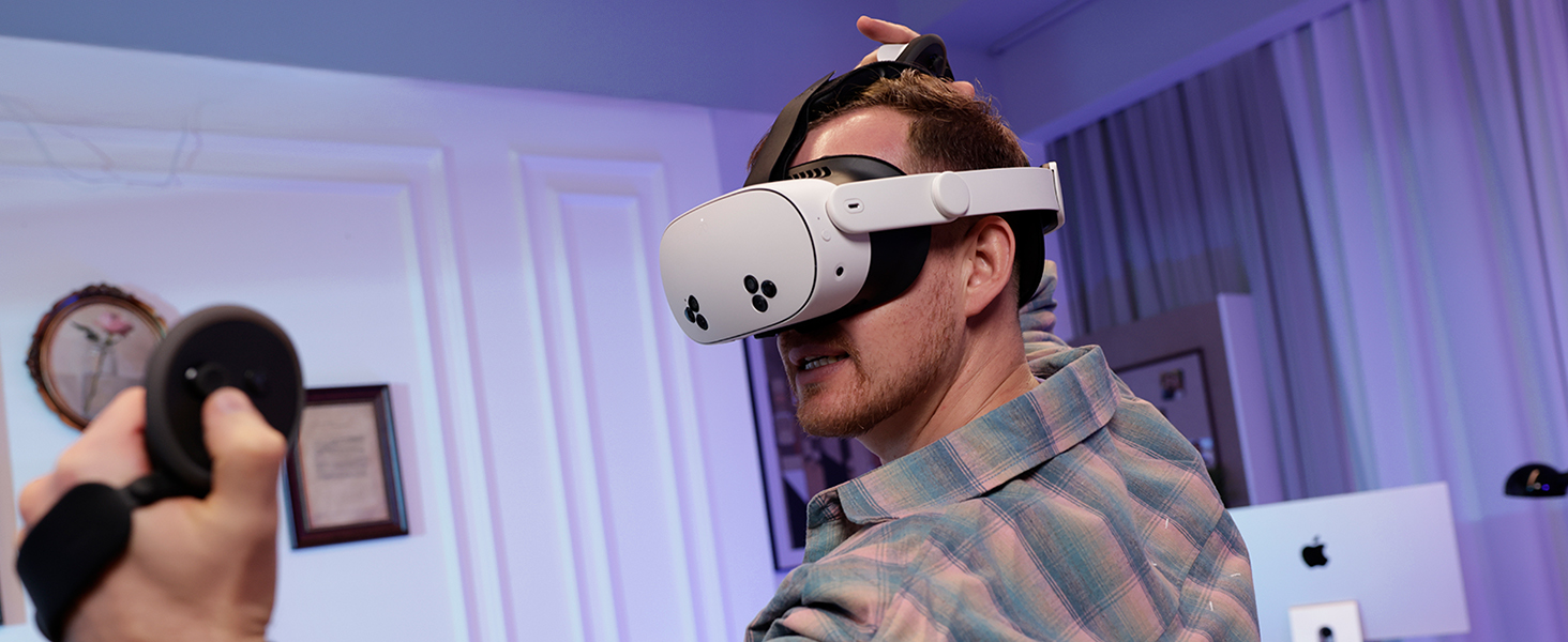 Man using virtual reality headset and controller with MacBook and framed picture in background.